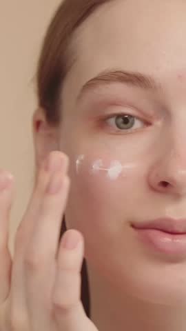 Young woman applying cream on her face close-up looking at camera, half face portrait, morning beauty routine, makeup preparation, skin care. Advertising concept. Vertical video