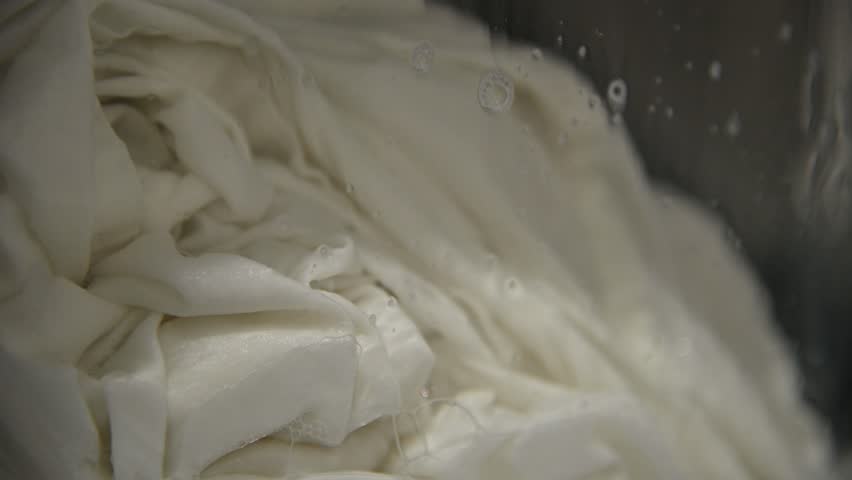 A close-up of old, yellowed gray laundry that was once white being washed in a washing machine. Bubbles of detergent drip down the glass window.