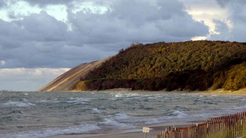 Lake Michigan’s powerful waves crash under a cloudy autumn sky, with Sleeping Bear Dunes rising in the background for a dramatic fall shoreline view wide shot stable.