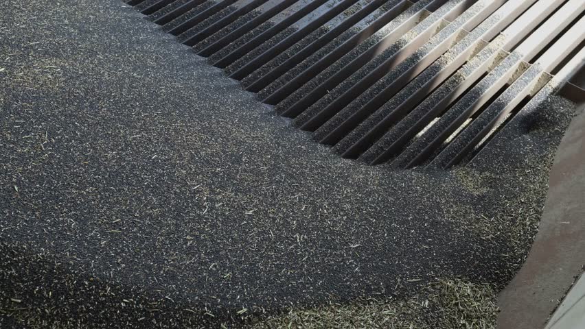 Slotted metal grate next to a mound of dark granular feed with pale fibrous bits in a processing facility, close up view with angled ribs and sunlight on edges