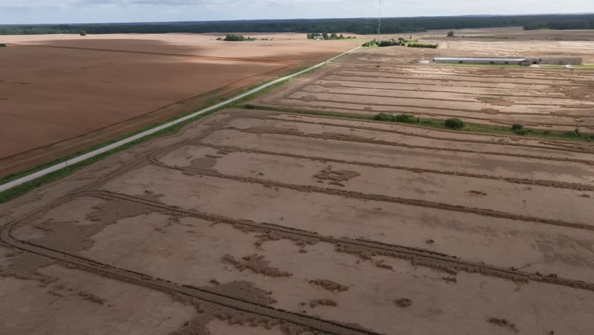 Aerial pan of harvested wheat strips, service road, mast, farm. Tractor tracks and stacked swaths show methodical harvest rows and subtle parallax across terrain.