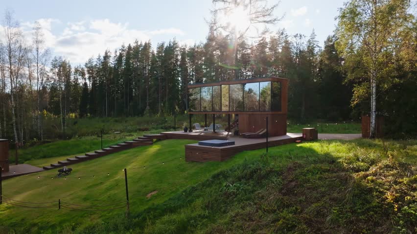 Modern glass cabin on a raised deck sits by birch and pine forest. Floor to ceiling windows mirror trees as sunburst and soft reflections sweep during slow push in.