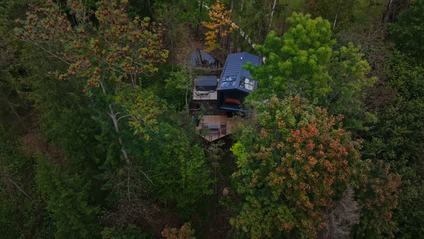 Aerial cabin with metal roof, glazed front, deck, hot tub, skylights. Drone drifts downward and rotates, revealing boardwalk, pond, autumn trees, warm interior lighting