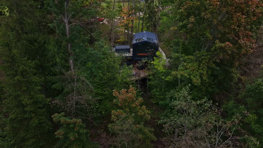 Aerial view of two story cabin in temperate woods, large glass front, skylights, L shaped deck, rooftop hot tub, pond and boardwalk, camera descends and pans
