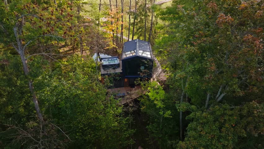 Aerial view shows modern two story cabin on elevated deck, glass wall, metal roof skylights, hot tub, boardwalk, and uphill pond in warm late afternoon light.