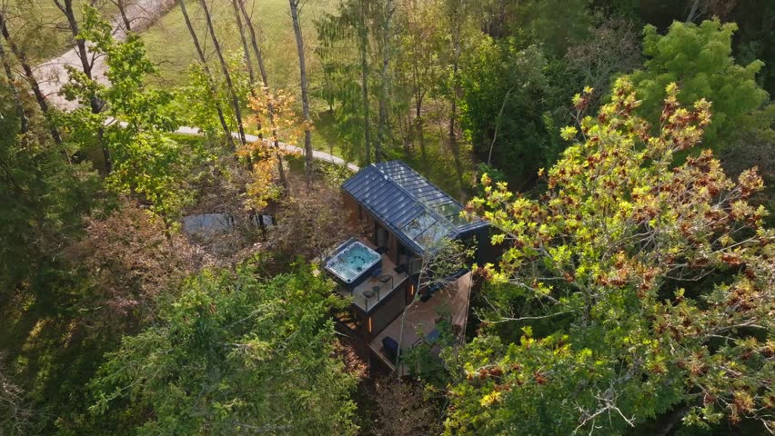 Aerial two story treehouse with rooftop glass panels at forest edge. Title in description. Camera pans and descends by paved walkway and meadow at late afternoon.