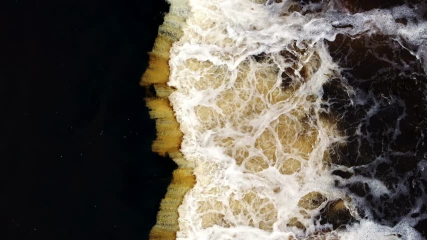 Aerial view shows dark water meeting a golden brown sandbar, with crashing waves and swirling foam. Title appears in description. Top down, strong contrast, rhythmic segments.