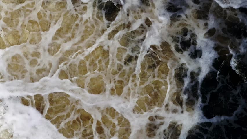 Aerial white swirls over golden brown water merging into near black. Title in description. Rapid turbulent motion in top down view with late afternoon side lighting.