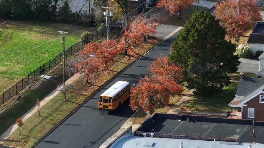 Aerial view of yellow school bus driving through an American residential neighborhood in autumn, with colorful fall trees, quiet streets and suburban houses under clear daylight. tracking shot.