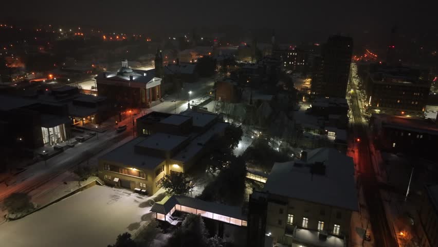 Drone flight over American city located on hill during snowy winter night. Lighting lantern along road with museum building and church tower. Snowy roofs and downtown of Lynchburg in back, Virginia