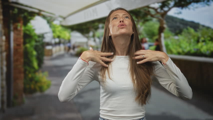 Woman with long hair and hands raised, face looking up in street gesturing near outdoor cafe canopy and walkway; frustration.