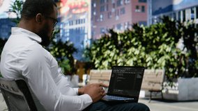 Confident adult reviews binary code script for his programming remote work, sitting on a bench in the city center and working on laptop. Scrolling and typing near lively downtown scenery. Camera B. - Powered by Shutterstock - Get 15% off with code: PIKWIZARD15