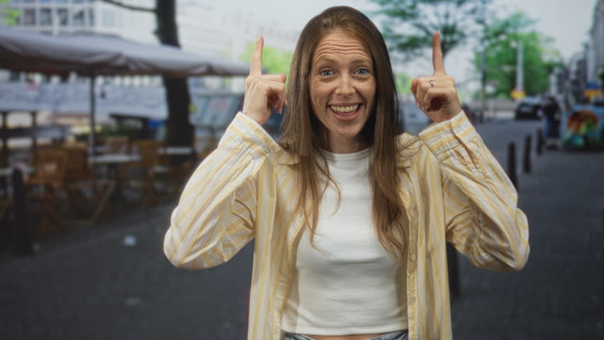 Young hispanic woman points both fingers up on a street terrace outside a cafe, smiling with eyes closed and laughing openly; joy.