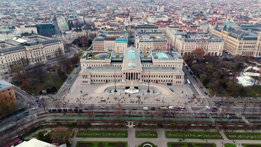 Aerial landscape drone shot panning over the Austrian Parliament Building, showing surrounding roads, moving traffic, and Vienna city blocks beneath clear winter sunlight and crisp seasonal shadows.