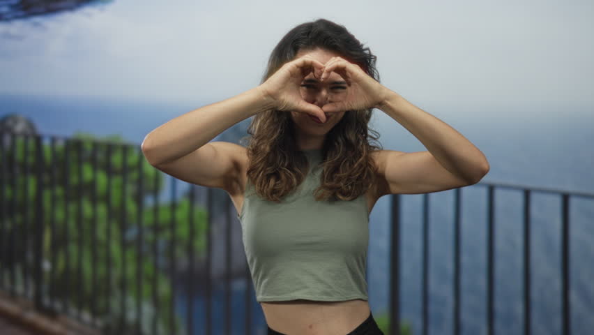 Woman with bare midriff smiling and forming heart shape with hands at building balcony; affection.