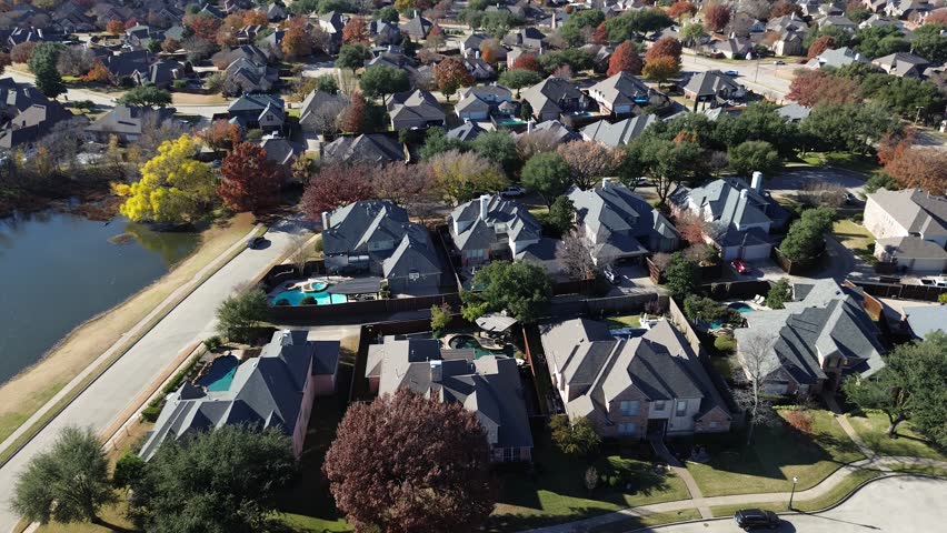 Two-story homes in Coppell, Texas arranged in grid pattern near scenic pond. Autumn foliage surrounds fenced yards and pools, blending natural and built environments in quiet suburban setting. USA