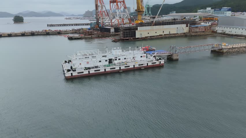 Aerial shot of an industrial port featuring a white work barge docked at a pier, alongside a busy shipyard with large cranes and ships under construction.