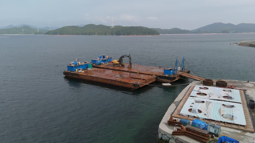 Aerial view of rusty industrial barges with an excavator docked at a concrete construction pier.