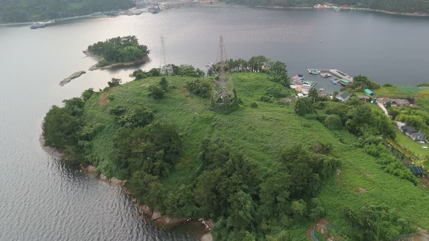 Panoramic aerial view of a lush green island with electricity towers, a coastal village, and aquaculture farms in serene waters, showcasing nature, infrastructure, and human life.