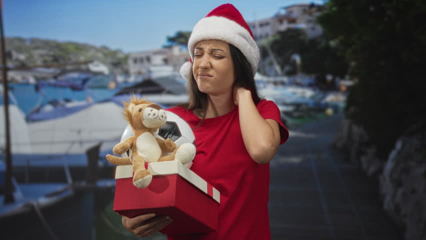 Woman holding red gift box with plush lion and soccer ball, hand on neck on a street near boats and marina; holiday fatigue.