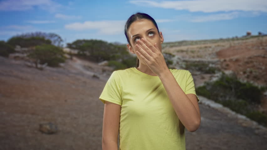 Young hispanic woman covering mouth with hand on forest path wearing yellow t shirt, eyes closed and leaning slightly forward; fatigue resignation.