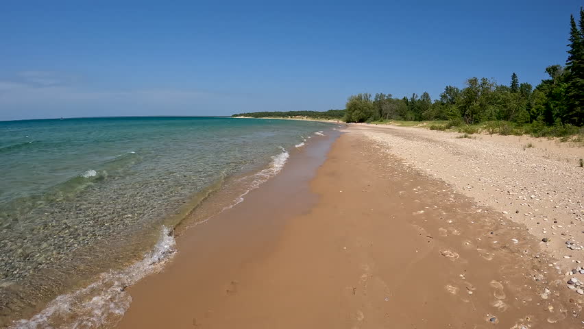 Stunning Natural Beauty on the Sandy Shore of Lake Michigan. Moving backwards along the coastline of Lake Michigan. Waves crash onto the sandy beach with rocks. The forest lines the beach. Unspoiled nature located within Wilderness State Park in Northern Michigan, USA.