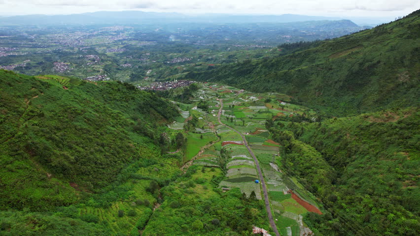 A small green “heaven valley” in the Dieng Plateau, Indonesia, known as the Swiss van Java, unfolds in cinematic aerial drone footage beneath a cool highland atmosphere. The valley is covered in lush vegetation, layered agricultural fields, and scattered rural settlements nestled among rolling hills. Narrow roads and pathways weave through the landscape, connecting the villages hidden within the greenery. Beyond the foreground valley, another vast valley opens dramatically into the distance, filled with clusters of villages and cultivated land stretching toward the towering slopes of Mount Sindoro. The volcano rises prominently on the horizon, partially wrapped in mist and cloud, dominating the highland scenery. The composition highlights volcanic geography, rural mountain life, and the breathtaking layered landscapes of Central Java in Southeast Asia from a sweeping aerial perspective.

