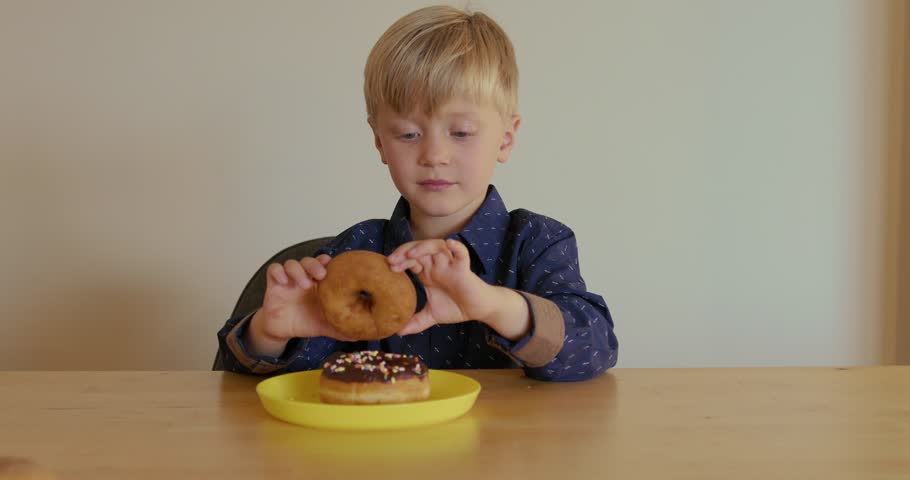 A joyful young boy with two chocolate donuts on a plate lifts one and looks through the hole of the donut. Enjoyment of sweets