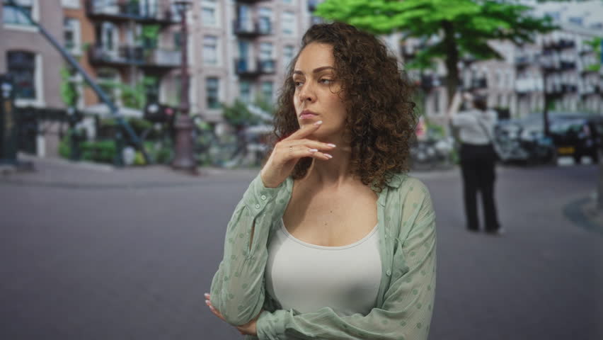 Woman hand on chin on city street wearing white tank top and sheer shirt showing chest; pensive reflection solitude.
