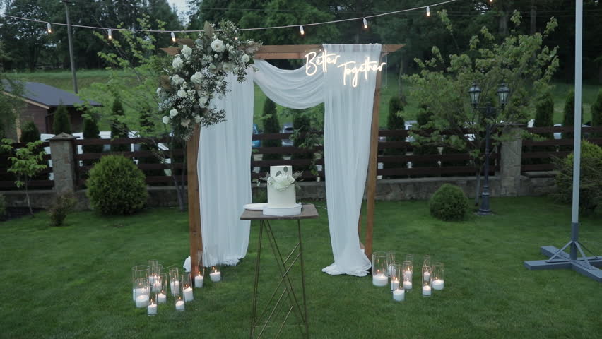 a stylish wooden wedding arch outdoor, decorated with white fabric, with a luxurious white wedding cake standing in the center.
