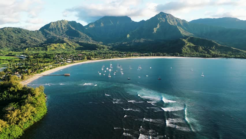 Aerial drone footage in 4K of Hanalei Bay on the island of Kauai Hawaii. Well known surf location and iconic tropical coastline featuring gentle waves, clear ocean water, and dramatic mountain backdrops.