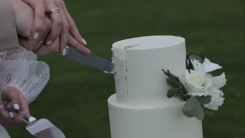 A close-up of the bride and groom holding the knife together as they cut a luxurious two-tier white cake decorated with flowers