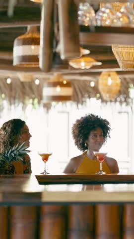 Multiethnic friends sitting at a bar counter, drinking colorful cocktails, and engaging in lively conversation in a stylish tropical resort. Vertical