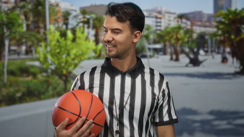Young hispanic man outdoors holding basketball wearing referee shirt in sunny urban background with palm trees and blurred cityscape expressing sports enthusiasm.