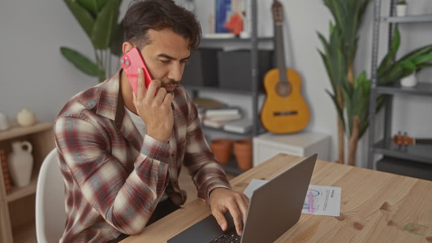 Young man in office using laptop and phone, surrounded by modern decor, showcasing a professional environment with personal touches like plants and a guitar.