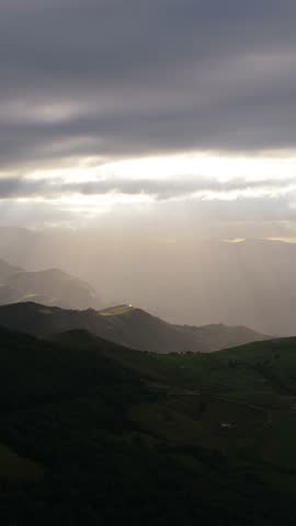 Majestic sunbeams breaking through dramatic dark clouds, illuminating a vast green mountain valley landscape during a beautiful sunrise. Vertical footage
