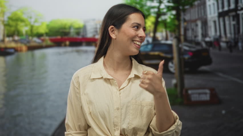 Woman pointing thumb to canal on a street in amsterdam, finger to lips for silence; playful travel.