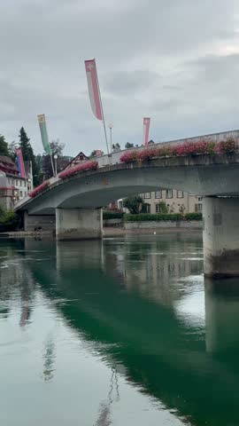Peaceful view of Stein am Rhein, Switzerland, on a rainy day. Features the Rhine bridge with Swiss flags, historic half-timbered houses, and emerald river reflections under a moody, overcast sky.