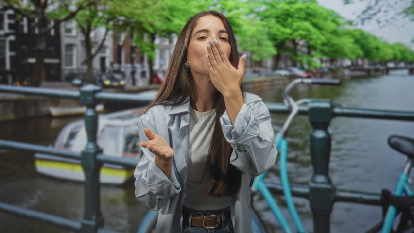 Woman blowing a kiss with hands cupped on a canal bridge in a street, wearing a light shirt and jeans while a bicycle leans nearby; playful travel moment.