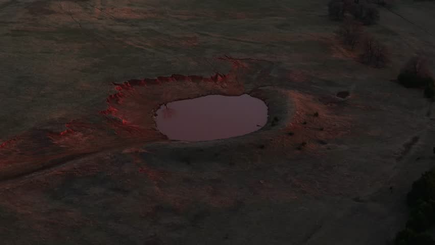 Aerial view of a small pond surrounded by red soil and grassland, showing natural erosion patterns and uneven terrain in a rural landscape under low-angle light.