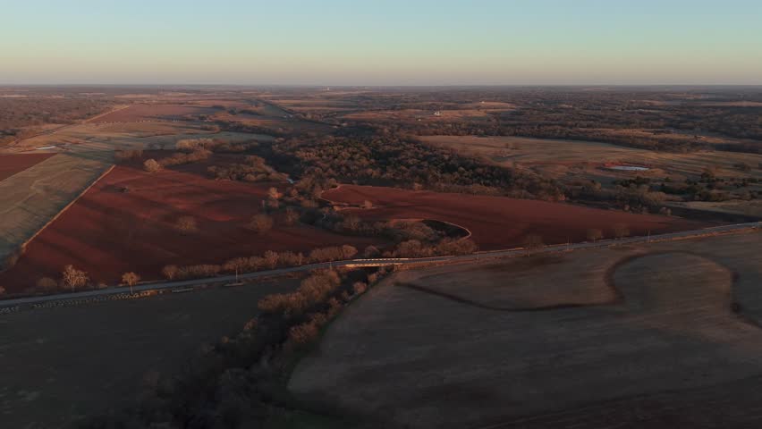 Wide aerial view of rural farmland with red soil fields, tree lines, and a country road cutting through rolling agricultural landscape under clear sky.