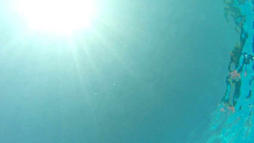 View from bottom of pool to sexy woman swimming under water with sun flare at background. Girl wearing red bikini floating in basin. Female tourist enjoying resting on resort. Vacation concept