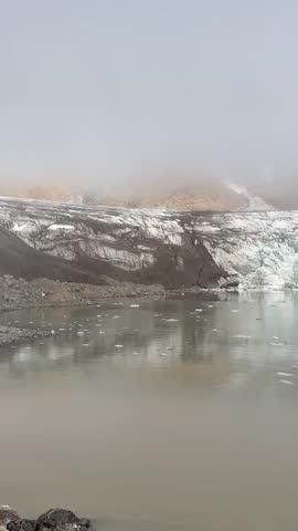 The Rasmussen Glacier stretches across a remote part of Greenland, surrounded by snow-covered mountains and Arctic terrain, with icy formations and glacial rivers visible in the landscape.