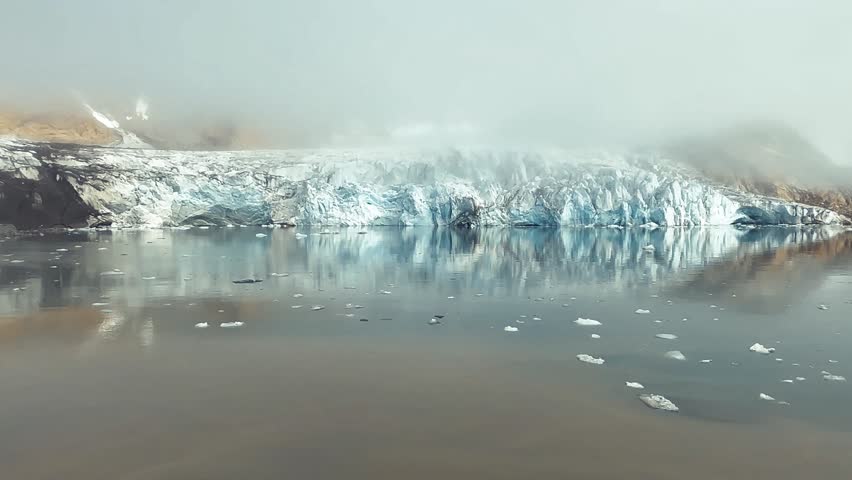 The Rasmussen Glacier stretches across a remote part of Greenland, surrounded by snow-covered mountains and Arctic terrain, with icy formations and glacial rivers visible in the landscape.