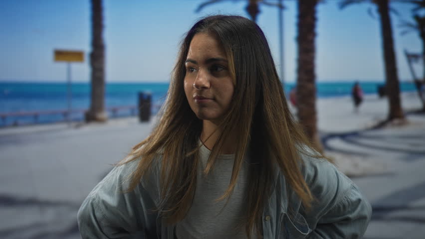 Young hispanic woman points finger toward camera on seaside promenade under palm trees; youth confidence.