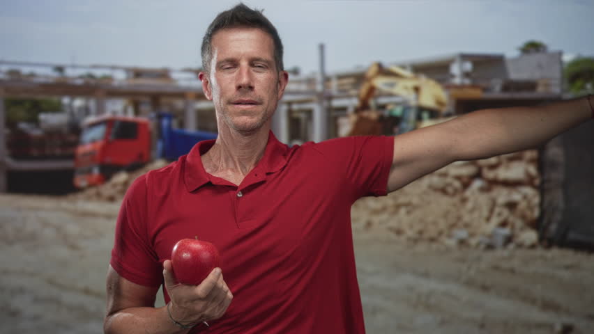 Man holding red apple, hand on cheek for toothache at construction building near excavator and truck; pain.