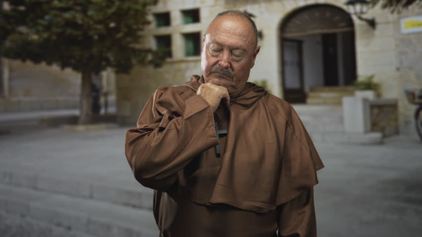 Man monk with hand on chin, eyes closed, standing before a stone church building in brown habit; quiet contemplation.