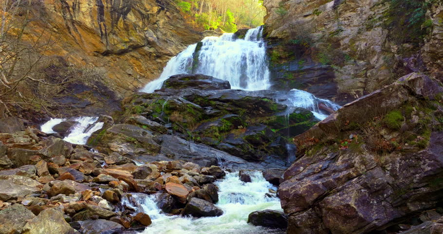 Cullasaja Falls in North Carolina with powerful water pouring through a narrow gorge. Autumn leaves create a golden canopy along the cliffs
