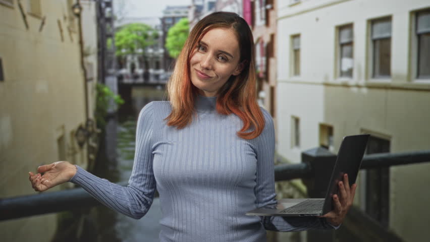 Woman holding laptop with palms up shrug on a street canal balcony, gentle smile, light blue sweater and red hair; curiosity freelance.