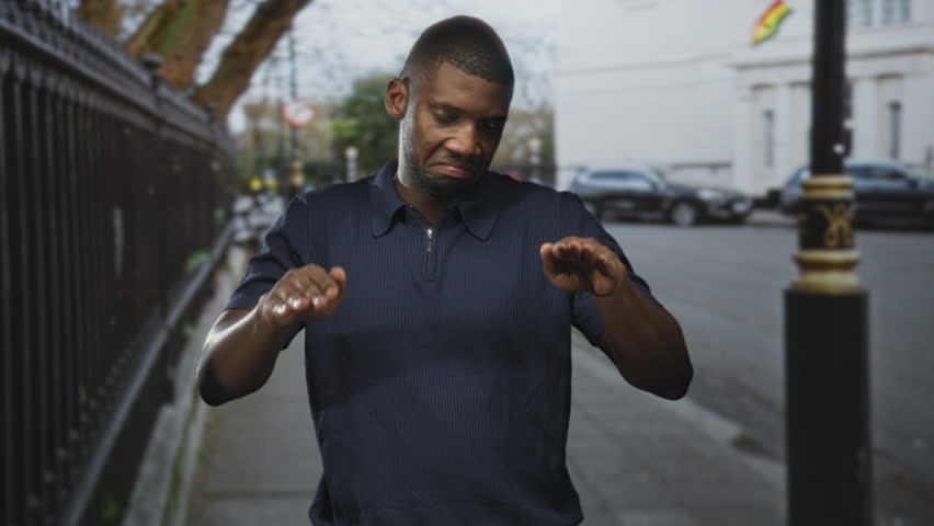 Man with palms forward gesture and extended hands as if pausing midstep on a paved street near cars, railing and a lamppost; hesitation.